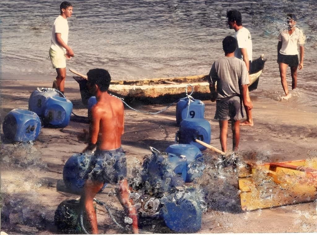 Fotografia antiga em praia do litoral, com canoa caiçara e bombonas na areia — cena da vida no litoral.